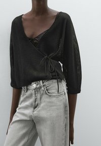 Schwarzes Wickeloberteil mit dreiviertellangen Ärmeln, strukturiertem Stoff, in der Taille gebunden. Kombiniert mit hellgrauen, hoch taillierten Jeans mit Knopfverschluss.