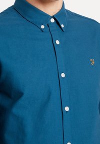 Camisa de botones de color verde azulado con cuello puntiagudo, con botones blancos y un pequeño logo bordado en oro en el pecho. Textura suave de algodón.