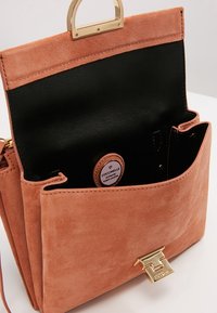 Torebka zamszowa w kolorze brązowym z złotym zapięciem, otwartą klapką i czarnym wnętrzem z widoczną metką marki Coccinelle.