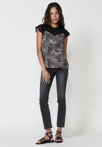 Top senza maniche nero e beige con dettagli in pizzo, abbinato a jeans aderenti grigi scuri. I sandali presentano cinturini e borchie su una suola piatta.