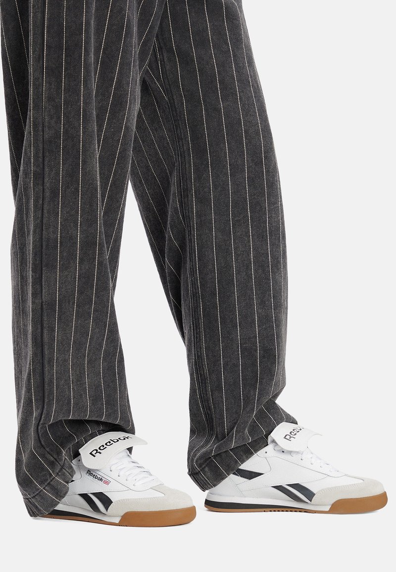 Baltas sporta apavi ar melniem akcentiem, mīkstu apkakli un gumijas zoli; valkāti ar pelēkiem svītrainiem platā stila biksēm.