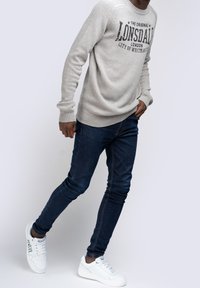 Light gray knit sweater with front logo, dark blue fitted jeans, and white sneakers with green accents. Casual, modern outfit.