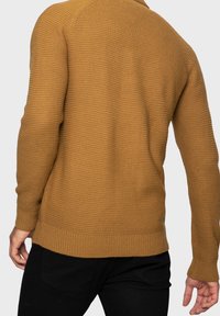 Sweter dziany w kolorze gorczycy z fakturowanym wzorem ściegu, długimi rękawami oraz ściągaczami na mankietach i dole, noszony na czarnych spodniach.