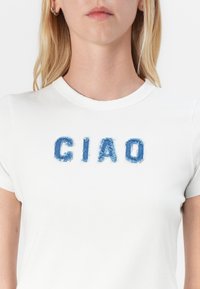 Hvit t-skjorte med rund hals som har blå, slitte denimbokstaver som staver "CIAO" på brystet. Korte ermer, myk bomullsstoff.