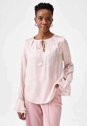 Kobieta ubrana w jasnoróżową bluzkę w groszki z falbanami na mankietach i wiązaniem przy dekolcie, w połączeniu z jasnoróżowymi, dopasowanymi spodniami, stojąca na białym tle.