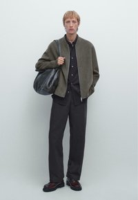 Giacca bomber grigia con zip, camicia nera con bottoni, pantaloni scuri e scarpe di pelle marroni. L'uomo porta una borsa nera.