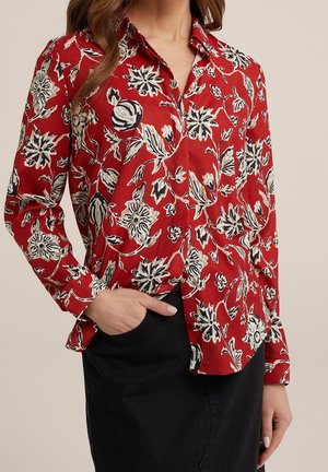 Woman wearing red long-sleeve shirt with white and black floral print, tucked into a black skirt, hand in pocket.