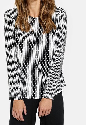 Femme portant un haut à manches longues noir et blanc à motifs rectangulaires verticaux, associé à un pantalon noir, debout avec les mains détendues.