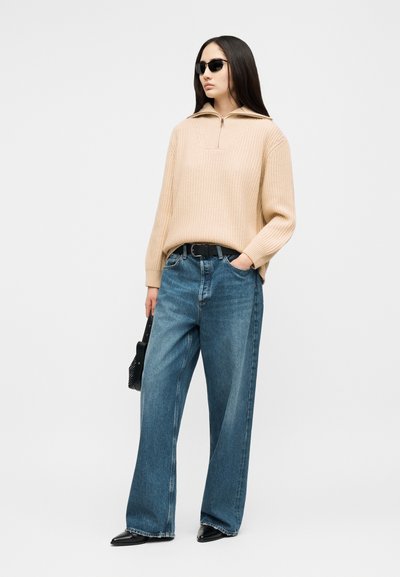 Femme portant un pull côtelé beige, un jean large bleu, une ceinture noire, des chaussures noires à bout pointu, des lunettes de soleil et tenant un sac à main noir.