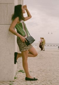 Top verde con estampado, falda de leopardo, zapatos planos negros y pequeño bolso cruzado negro, con un fondo de adoquines y cielo nublado.