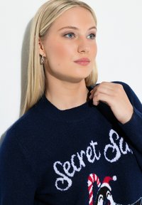 Młoda kobieta z blond włosami, nosząca srebrne kolczyki w kształcie kół i granatowy sweter świąteczny z napisem "Secret Santa" oraz wzorem w cukierka.
