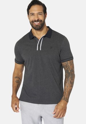 Lächelnder Mann mit dunklen Haaren und Bart, der ein dunkelgraues Poloshirt mit schwarzem Kragen und hellgraue Hose trägt und Tätowierungen auf beiden Armen zeigt.