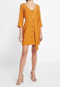 Senapsgul klänning med V-hals, 3/4-långa ärmar, knäppning fram och ett matchande bälte. Slätt tyg med en texturerad yta.