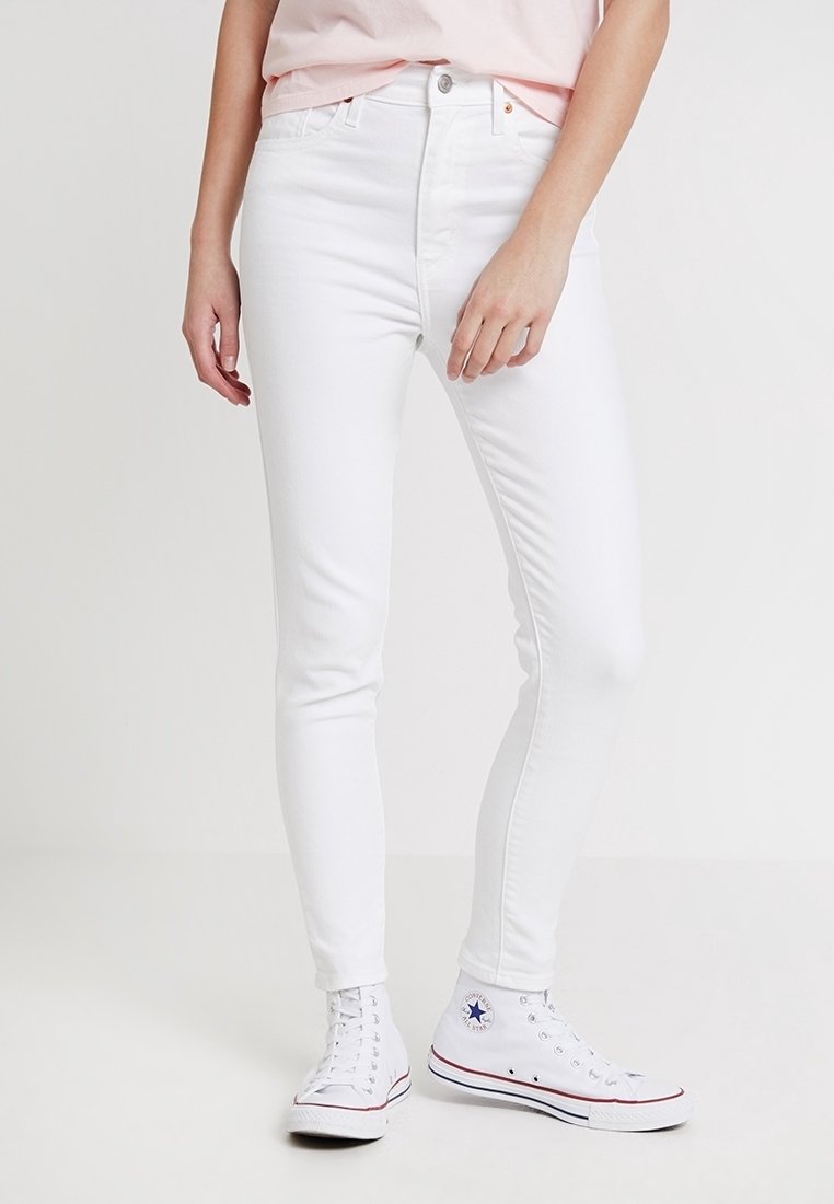 Personne portant un jean blanc slim, une chemise rose clair et des baskets blanches montantes, debout sur un fond clair uni.
