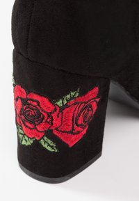Bloc-heel en daim noir avec des roses rouges brodées et des détails de feuilles vertes sur le côté extérieur du talon.