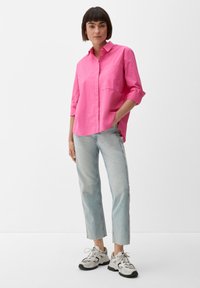 Roze overhemd met knopen, een borstzak, opgerolde mouwen en een ontspannen pasvorm, gecombineerd met lichtblauwe jeans en grijze sneakers.