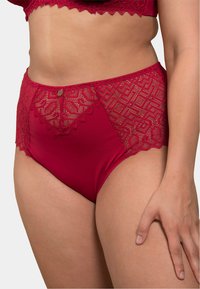 Culotte rouge taille haute avec des panneaux latéraux en dentelle présentant des motifs géométriques. Détail décoratif d'un bouton doré à l'avant qui rehausse le design.