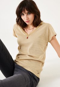 Camiseta de algodón beige con escote en V y mangas cortas, de tejido texturizado y cuello acanalado, combinada con jeans oscuros. Diseño simple sin patrones adicionales.