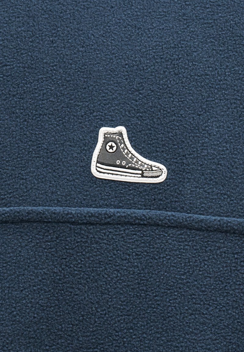 Dark blue textured fabric with a small patch of a black and white high-top sneaker sewn near a horizontal seam.