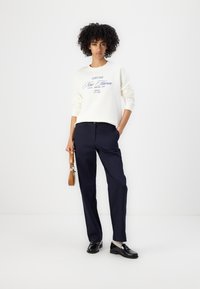 Pull léger beige avec texte bleu, coupe décontractée ; associé à un pantalon tailleur bleu marine et des mocassins noirs. Petit sac à main marron.