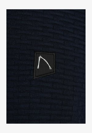 Tamsiai mėlynų mezginių audinys su tekstūruotu raštu, kuriame yra juodas stačiakampis pleistras su baltu geometriniu raštu.