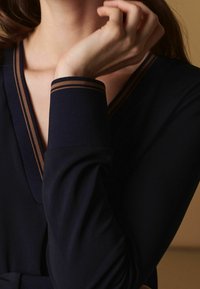 Maglietta a maniche lunghe blu navy con scollatura a V profonda e dettagli in marrone. Il tessuto morbido presenta un design aderente con una texture liscia.