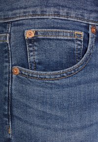 Blå denimjeans med en framficka, kopparnitar, kontrasterande gyllene sömmar och en mjuk, något sliten textur.