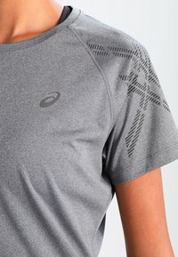 Camiseta deportiva gris de manga corta con una textura suave, que presenta un logo en el pecho y un detalle con patrón en el hombro.