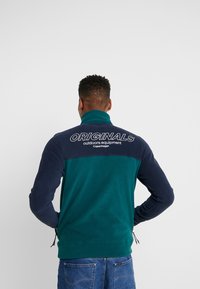 Giacca in pile color teal e blu navy, con collo alto e la scritta "ORIGINALS" stampata sulla schiena in bianco. Presenta polsini elasticizzati e orlo con coulisse.