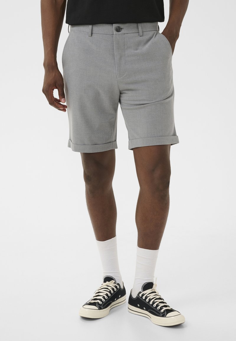 Homme portant un short gris clair à revers, des chaussettes blanches montantes et des baskets noires basses, debout avec les mains détendues sur les côtés.