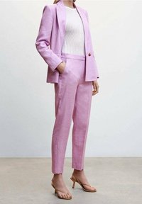 Frau in einem helllila Anzug mit maßgeschneiderter Hose und Blazer über einem weißen Rippenoberteil, kombiniert mit sandfarbenen High Heels.