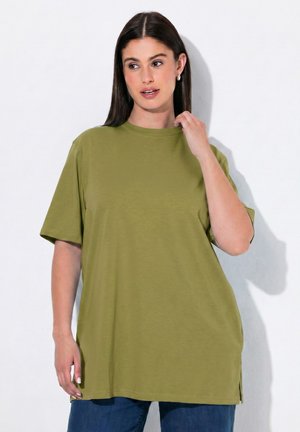 Femme aux longs cheveux foncés portant un t-shirt oversize vert olive et un jean bleu, touchant l'encolure, debout sur un fond blanc.