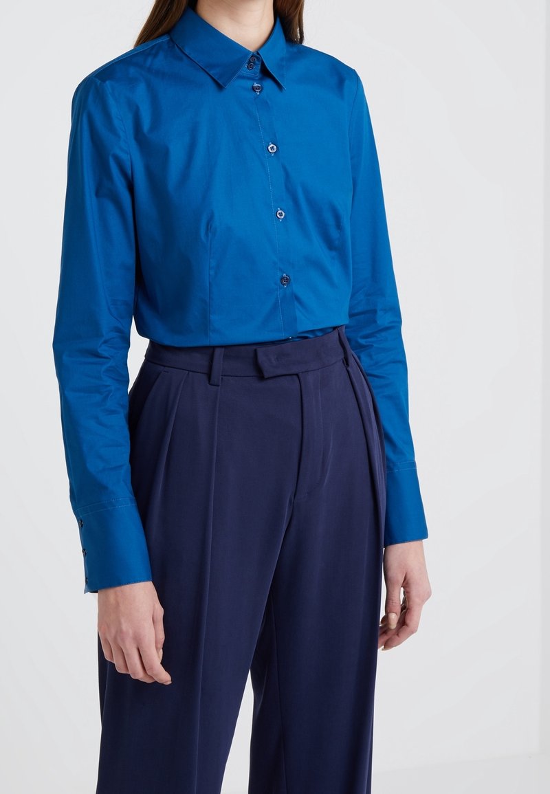 Personne portant une chemise boutonnée à manches longues bleu vif rentrée dans un pantalon plissé bleu foncé à taille haute.