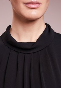 Svart blus med en vikt krage, veckdetaljer fram och ett mjukt, texturerat tyg. Närbild på halsringningen.