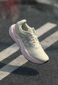 Beige og rosa løpesko med "FuelCell" såle i luften over asfalt med hvite linjer, som viser bevegelse og grep.