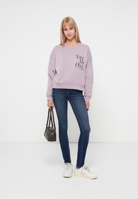 Jasnofioletowa bluza z czarnym napisem, zestawiona z ciemnoniebieskimi dżinsami skinny i białymi sneakerami. Modelka trzyma małą, ciemną torebkę.