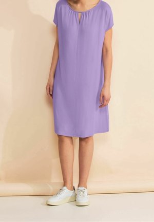 Femme portant une robe lavande courte aux genoux à manches courtes, associée à des baskets blanches, debout devant un fond beige.