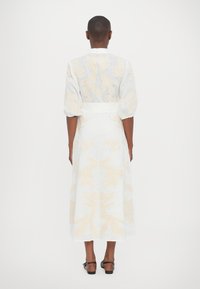 White linen dress with beige floral embroidery, long sleeves, and a fitted waist, reaching mid-calf length. Open back design.