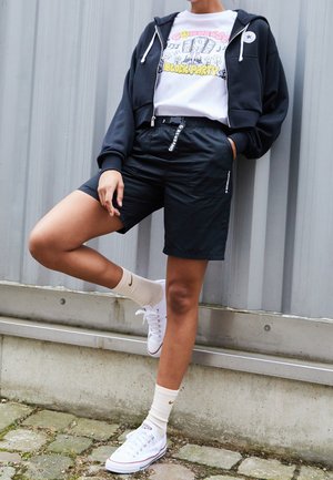 Person trägt schwarzen Kapuzenpullover mit Reißverschluss, weißes Grafik-T-Shirt, schwarze Shorts, weiße Socken mit Logo und weiße Sneaker und steht vor einer Wellblechwand.
