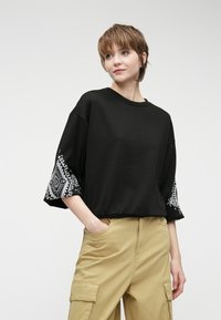 Melnas kokvilnas T-krekls ar trim ceturtdaļām, kuram ir balts ģeometrisks raksts uz piedurknēm, savienots ar khaki krāsas cargo biksēm.