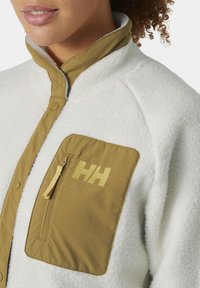 Valge fliistekiga jakk, kus on khaki värvi lukuga tasku, millel on kollane logo. Jakil on kõrge krae ja nööbiga kinnitamine.