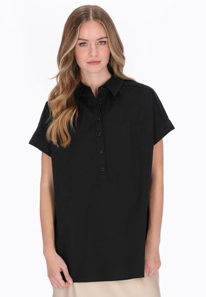 Młoda kobieta z długimi, falującymi blond włosami, ubrana w luźną czarną koszulę z krótkim rękawem i guzikami, stojąca na tle białego tła.
