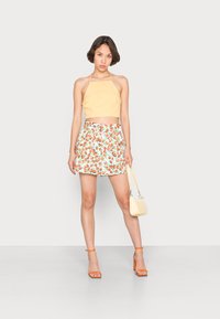 Top crop amarelo, calções brancos com padrão de frutas vermelhas e verdes, sandálias de salto laranja, mala amarela pequena, tecido suave, design ajustado.