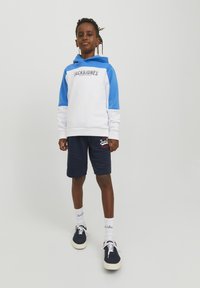 Valkoinen ja sininen huppari etulogon kanssa, yhdistettynä tummansinisiin shortseihin. Varustettu tummansinisillä kengillä ja valkoisilla sukilla, joissa on logotyyppinen yksityiskohta.