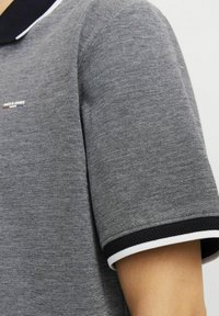 Pilkas polo marškinėlis su tekstūruotu audiniu, juodu apykakle ir kontrastinėmis baltomis detalėmis ant rankovių. Diskretiškas logotipo elementas.