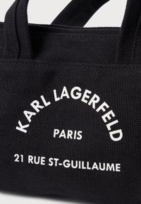 Μαύρη υφασμάτινη τσάντα ώμου με λευκό κείμενο που γράφει "KARL LAGERFELD PARIS 21 RUE ST-GUILLAUME" και δύο ανθεκτικές λαβές.