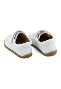 Zapatitos blancos para bebés con suelas de goma marrón, correas de Velcro y perforaciones decorativas, mostrados desde la parte trasera sobre un fondo blanco.