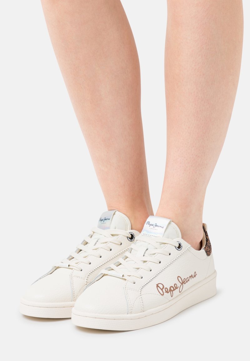 Baskets blanches Pepe Jeans avec un logo marron sur les côtés portées sur des jambes nues sur un fond blanc.