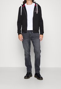 Persona con sudadera negra con cremallera y cordones blancos, camiseta blanca, jeans grises y botas negras con cordones, de pie contra un fondo liso.