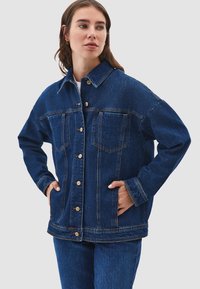 Kvinna i mörkblå denimjacka och jeans, med händerna i jackfickorna, tittar åt sidan mot en enkel grå bakgrund.
